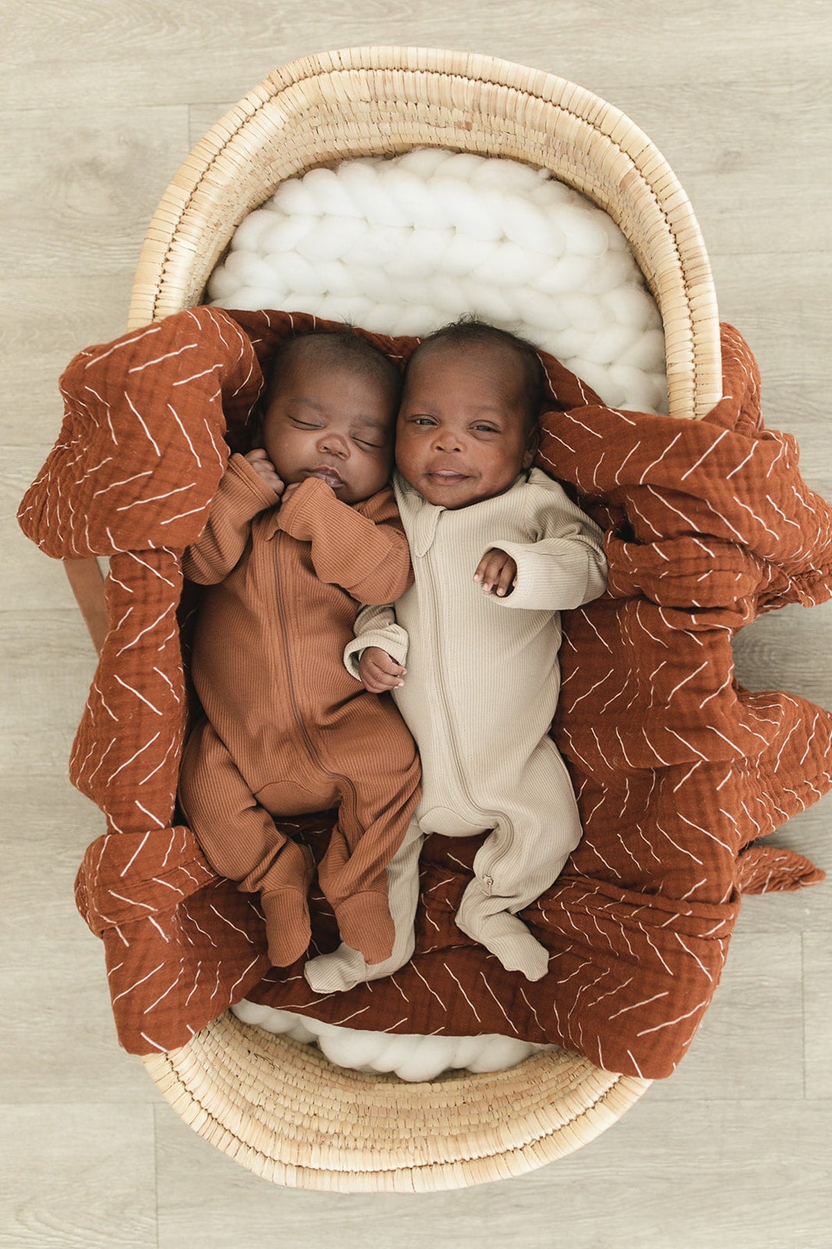 Two infants lie side by side in a woven bassinet, one wearing a brown ribbed footed pajama and the other.