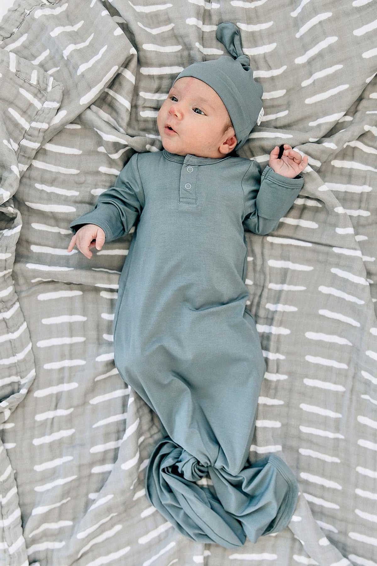 A baby in a dusty blue bamboo knot gown and hat lies on a patterned gray blanket.