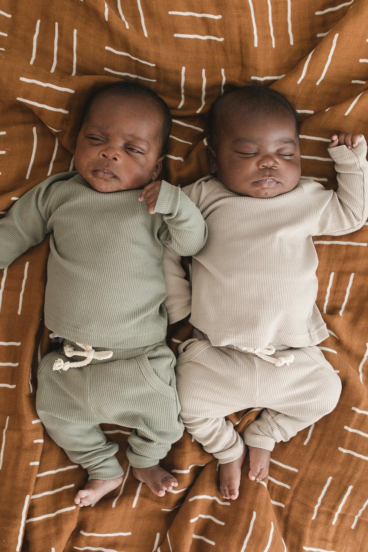 Two babies in ribbed outfits lie on a mustard mudcloth quilt with a geometric pattern.