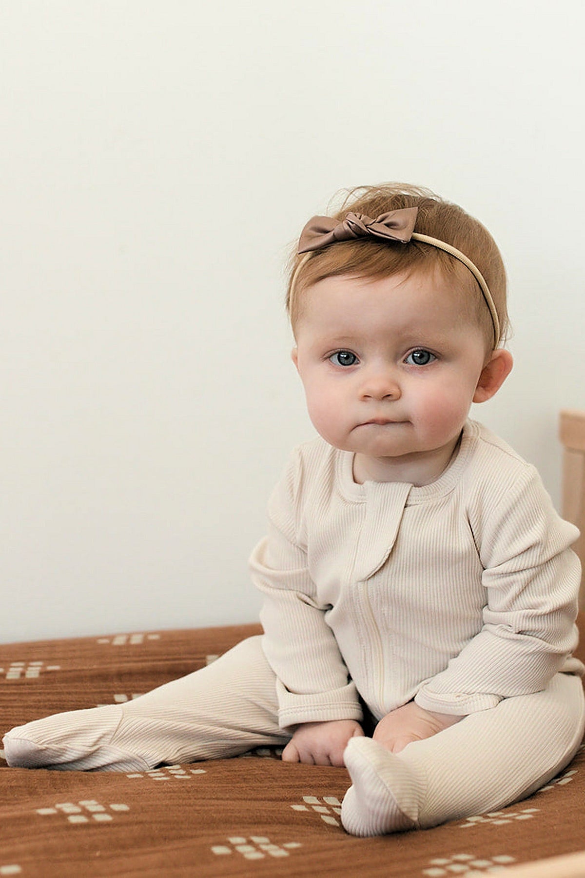 A baby sits on a brown patterned blanket, wearing a beige ribbed pajama with a zipper and a matching headband.