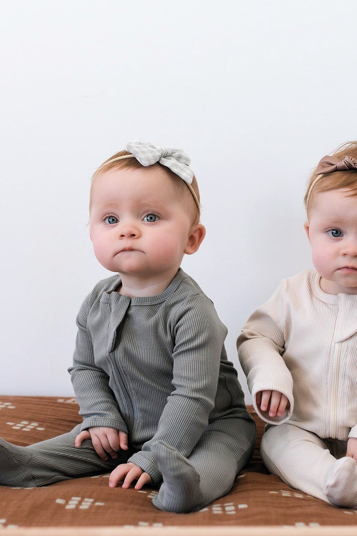 Two babies sit wearing ribbed footed pajamas; one in grey, the other in cream, both with headbands.