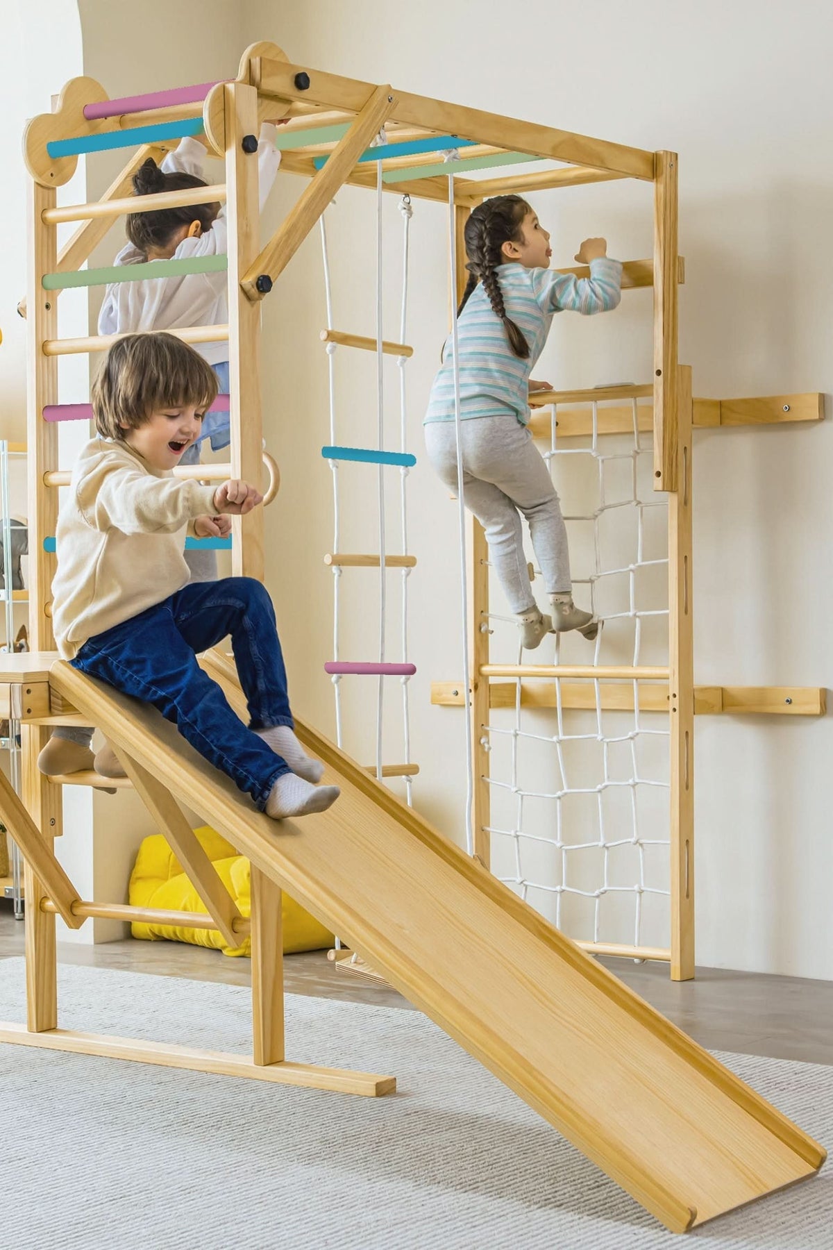 Three children play on a wooden indoor jungle gym, featuring a slide, climbing net, and colorful bars. The structure is designed for safe, active play in a modern setting.