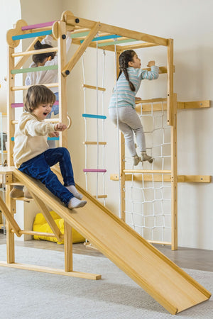 Three children play on a wooden indoor jungle gym, featuring a slide, climbing net, and colorful bars. The structure is designed for safe, active play in a modern setting.