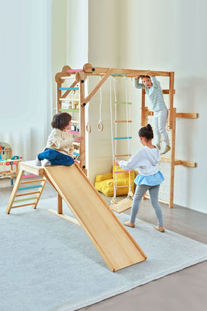 Children play on the Grove Foldable Jungle Gym Playset, featuring a wooden slide, climbing bars, and rings. The colorful design adds a vibrant touch to the indoor play space.
