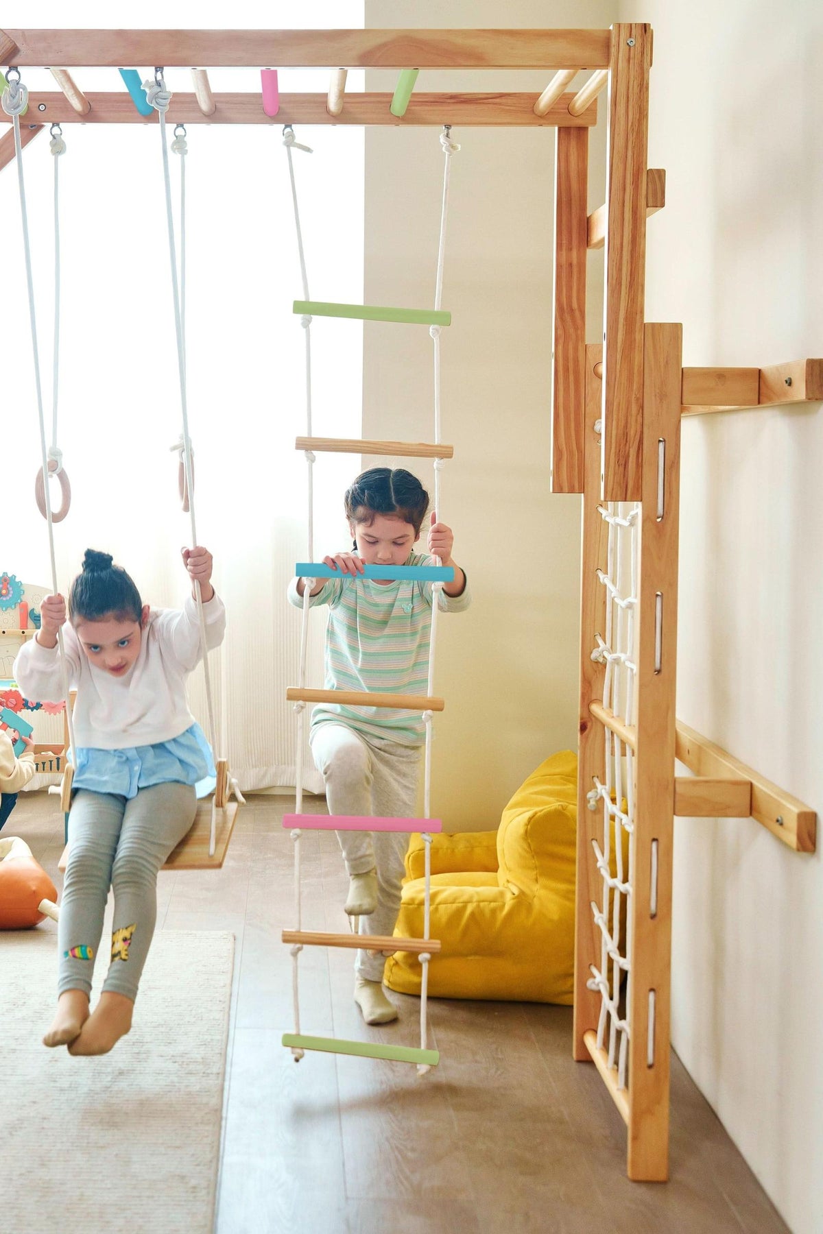 Two children play on a wooden jungle gym with swings and a climbing ladder. The gym features a natural finish, colorful bars, and is designed for indoor use.