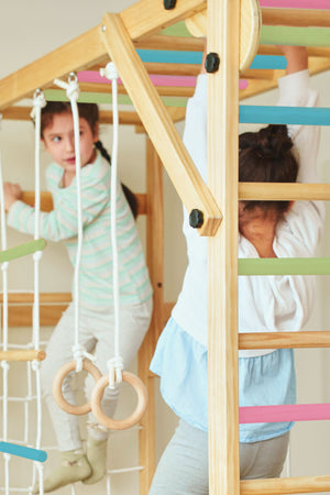 Two children play on a wooden foldable jungle gym featuring colorful bars and climbing elements. The structure is designed for indoor use, promoting active play and coordination.