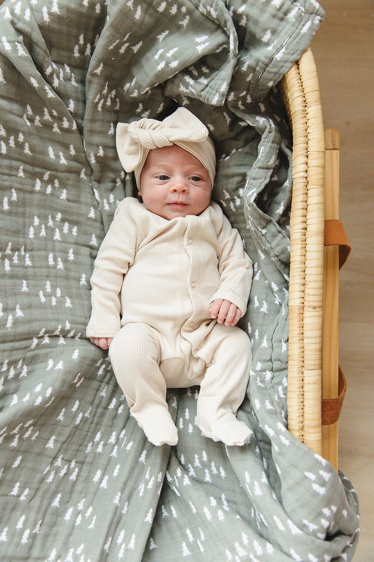 A baby in vanilla ribbed pajamas with a matching headband lies on a green patterned blanket in a basket.