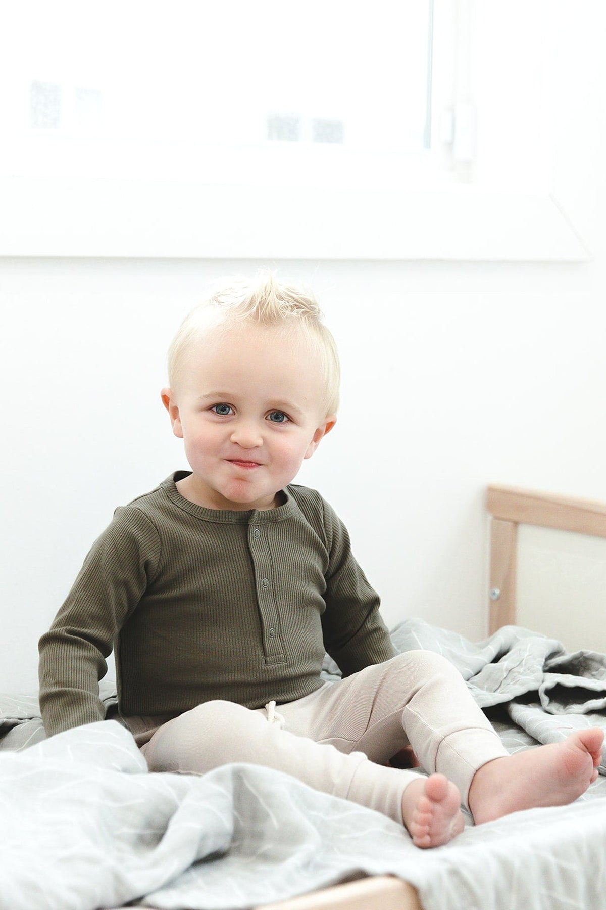 A toddler wearing a winter green ribbed long-sleeve bodysuit sits on a bed with light-colored bedding.