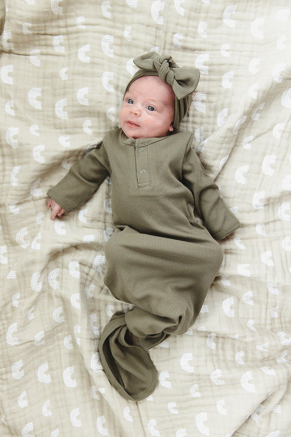 A baby wearing a green ribbed knot gown and matching headband lies on a patterned beige blanket.