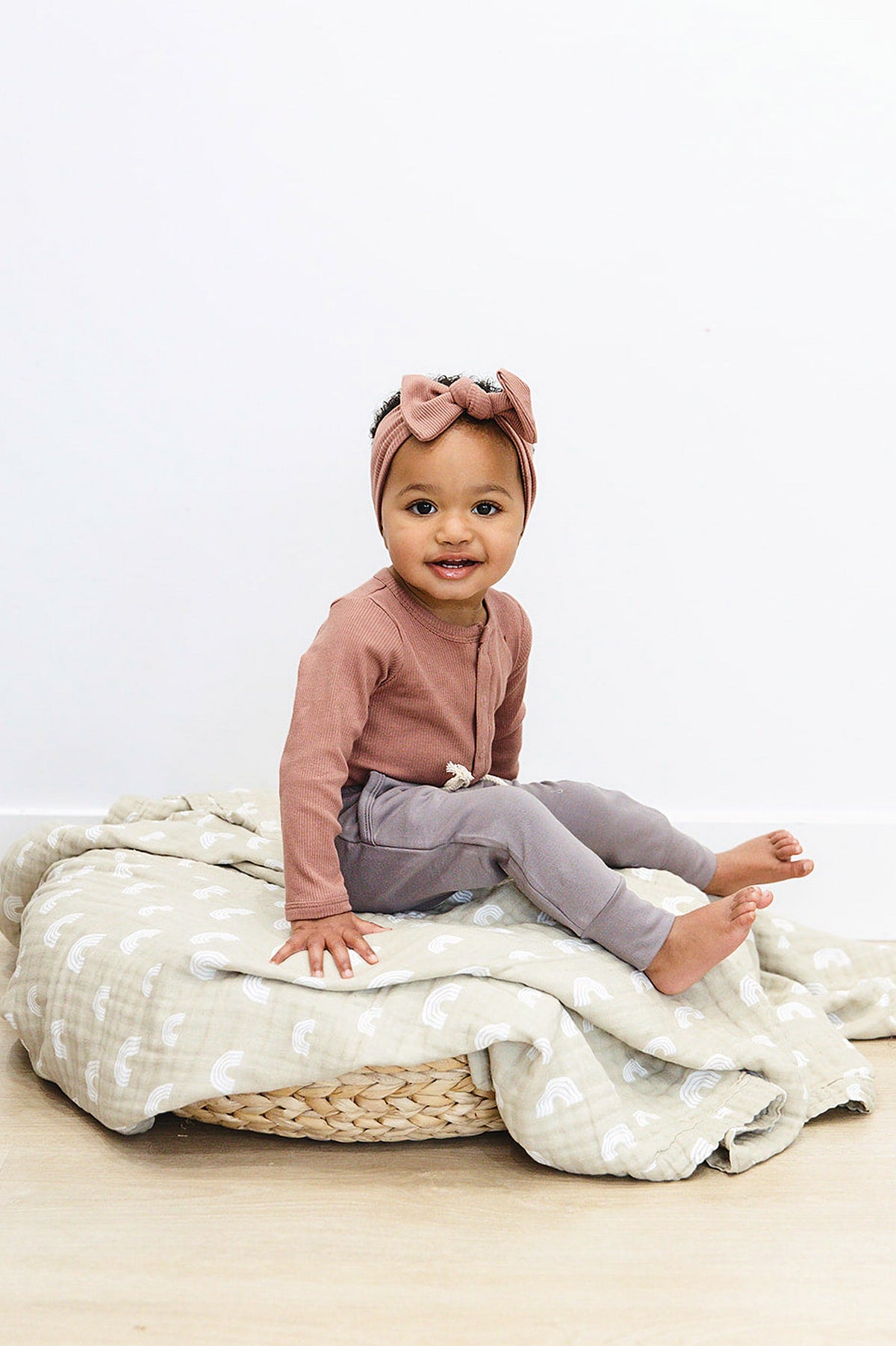 A baby wearing a dusty rose long sleeve bodysuit and matching headband sits on a patterned blanket.