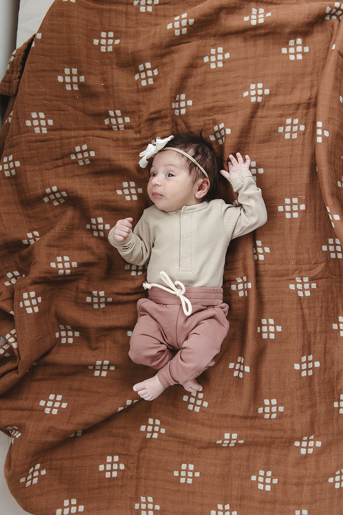 A baby wearing a beige ribbed long sleeve bodysuit and pink pants lies on a brown patterned blanket.