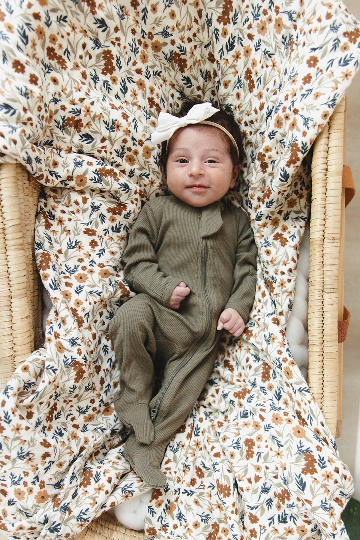 A baby in a green ribbed pajama lies on a floral-patterned blanket, wearing a white bow headband.