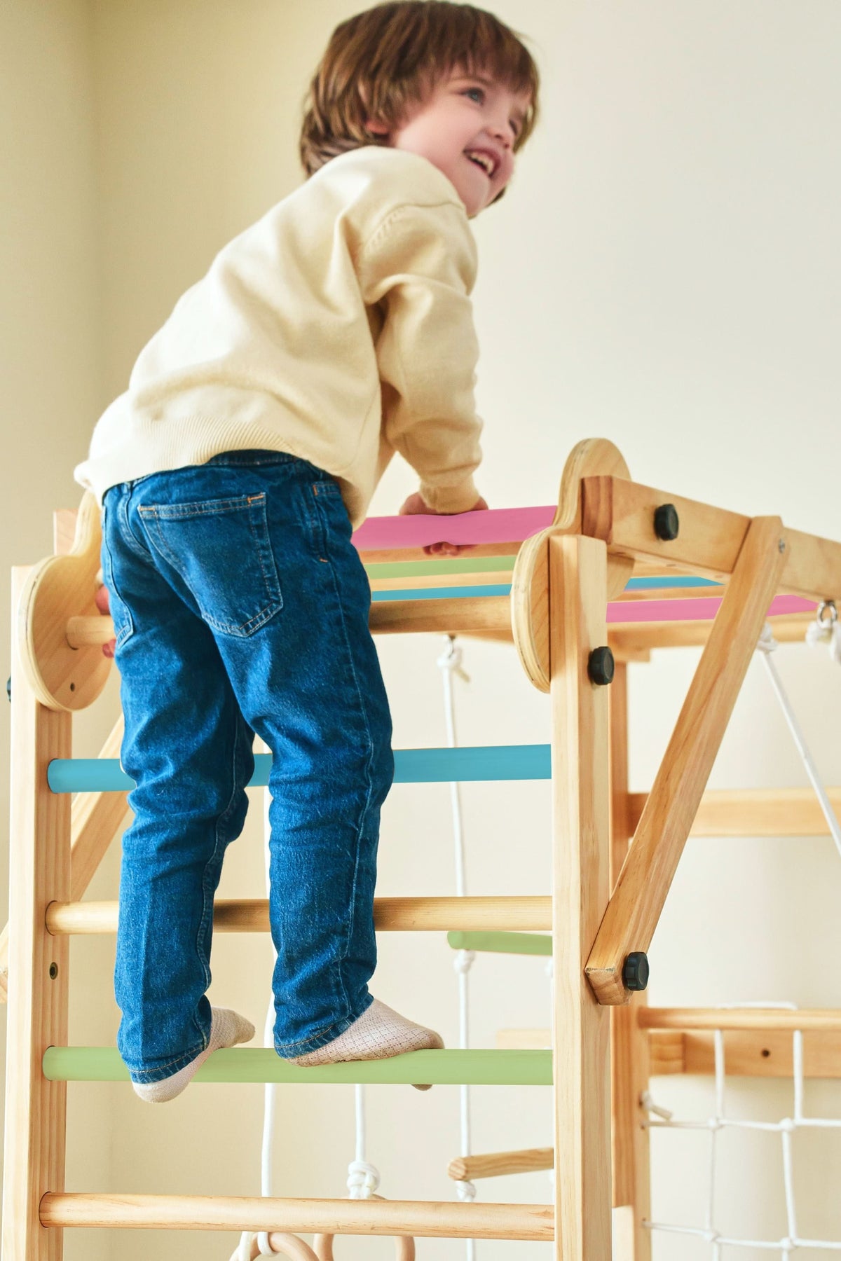 A child climbs the colorful rungs of a wooden jungle gym playset. The smiling boy wears a beige sweater and blue jeans, showcasing a safe and engaging indoor play environment.