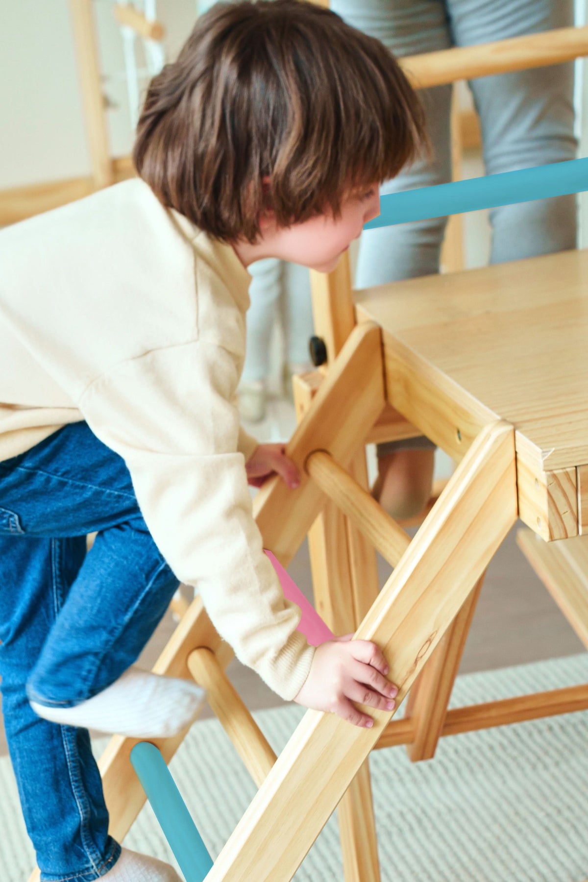 A child wearing a beige sweater and blue jeans climbs a wooden ladder with colorful bars on a foldable jungle gym playset, designed for indoor play.
