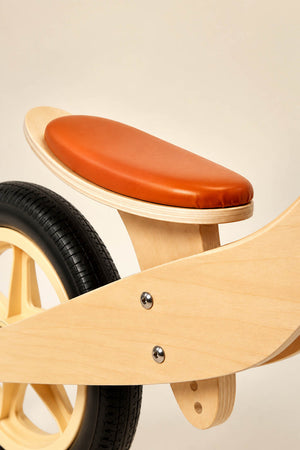 Close-up of the Maxi Wooden Balance Bike featuring a natural wood frame and a stylish brown leatherette seat. Designed for children ages 3 to 5, it emphasizes safety and balance.