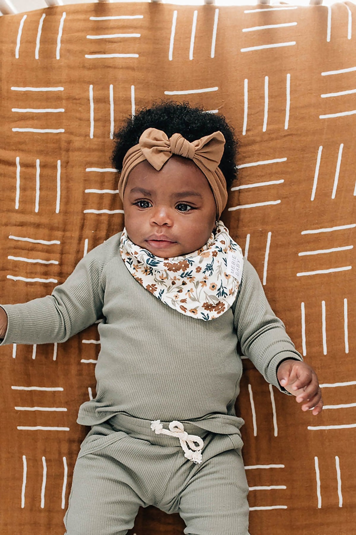 Baby wearing a floral muslin bib and green outfit, lying on a brown patterned blanket with a matching headband.