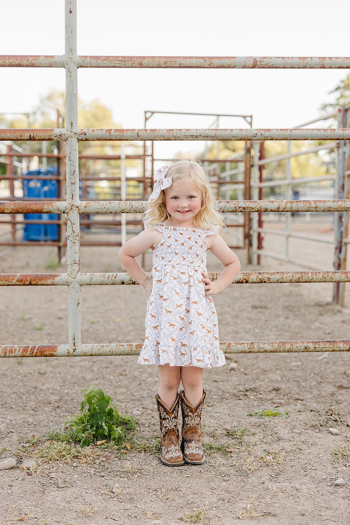 A young girl stands confidently in a pony-printed smocked dress, paired with brown cowboy boots, in a rustic outdoor setting surrounded by a metal fence and dirt ground.
