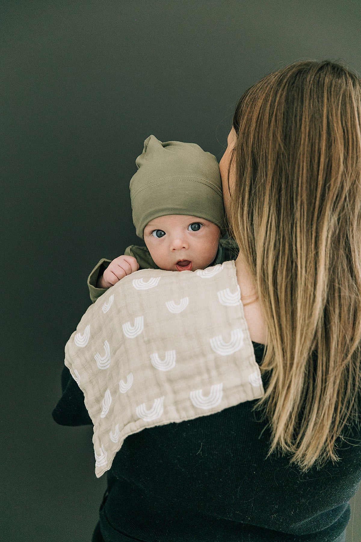 A baby wearing an olive knot hat is held by a woman with a patterned cloth over her shoulder.