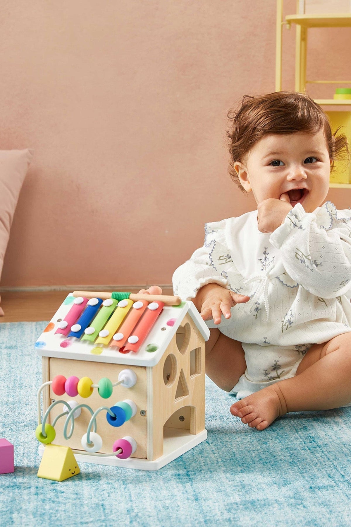 A toddler sits on a blue rug, engaging with a colorful wooden activity cube featuring various interactive elements.