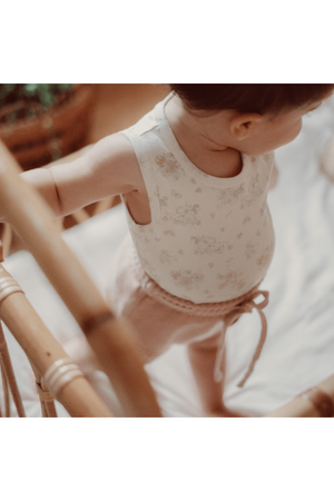 A baby stands in a cozy crib, wearing a floral-printed cotton tank top and soft pink shorts.