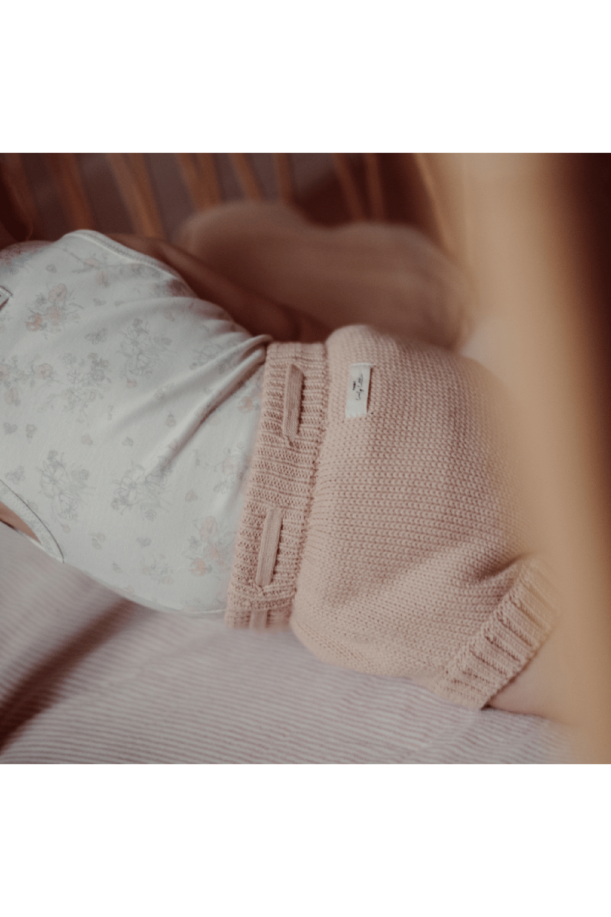 A baby is resting in a crib, wearing a floral-patterned onesie and soft, knitted shorts in a light pink color. The cozy setting features warm.