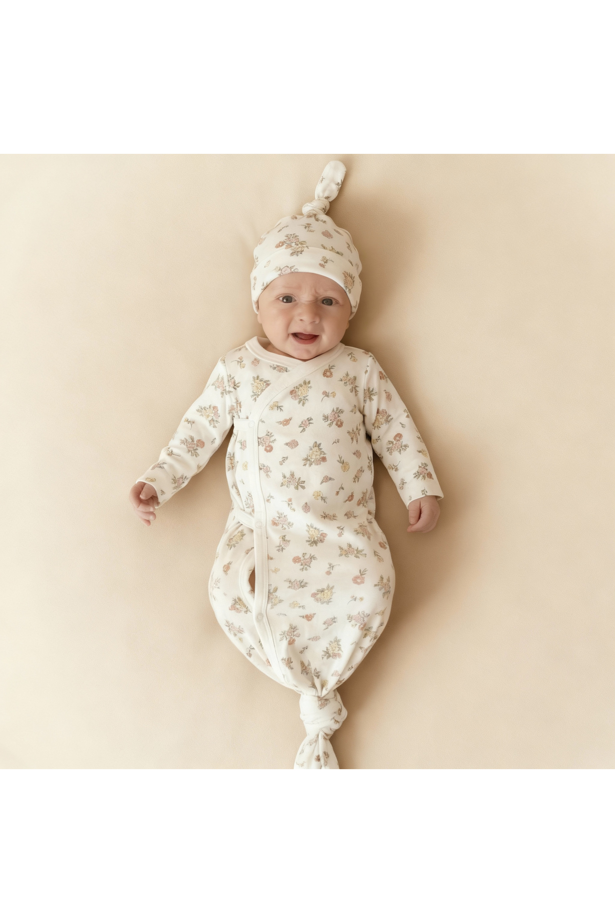 A smiling baby wears a soft, floral-patterned kimono knotted sleep gown and matching hat, both crafted from organic cotton. The light background accentuates the gentle design and colors.