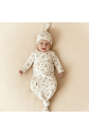 A smiling baby wears a soft, floral-patterned kimono knotted sleep gown and matching hat, both crafted from organic cotton. The light background accentuates the gentle design and colors.