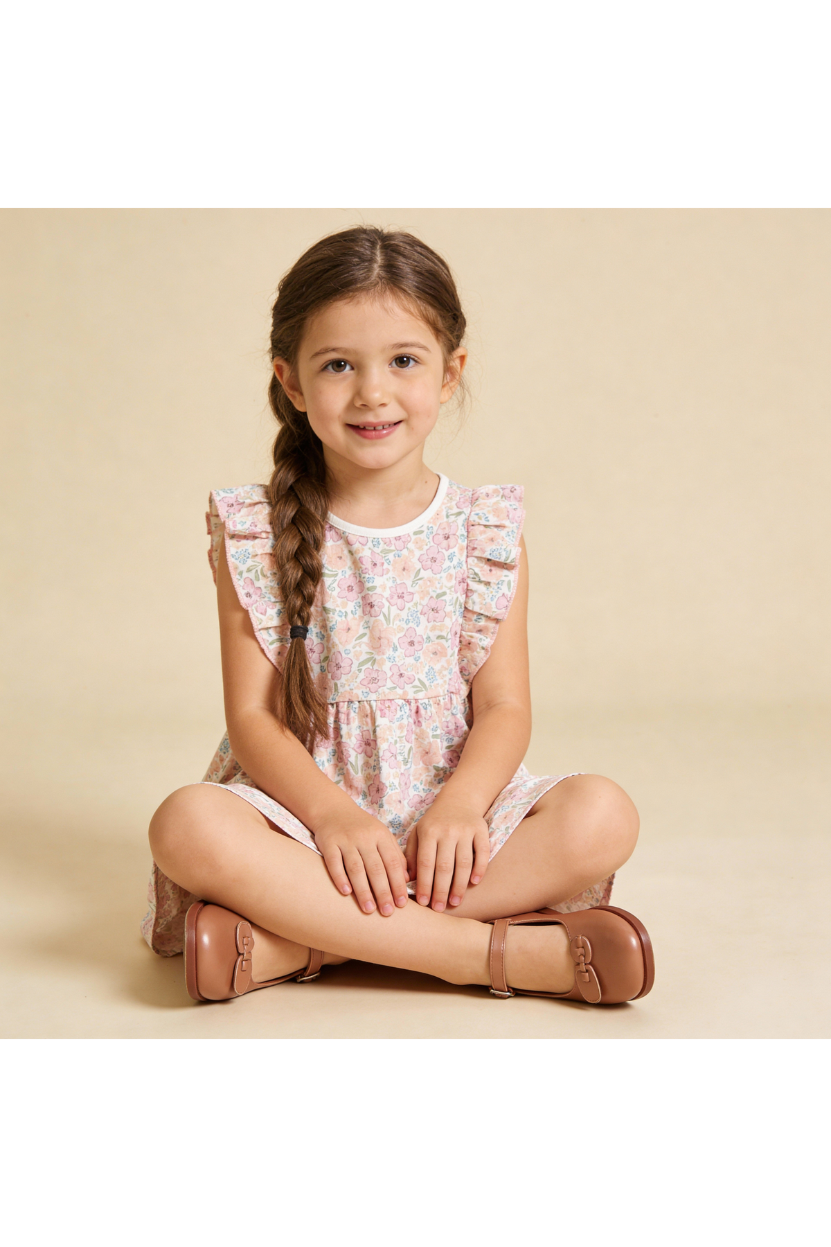 A young girl sits cross-legged on a neutral background, wearing a floral flutter dress with ruffled sleeves. Her brown shoes complement the playful design, showcasing a joyful, organic cotton outfit.