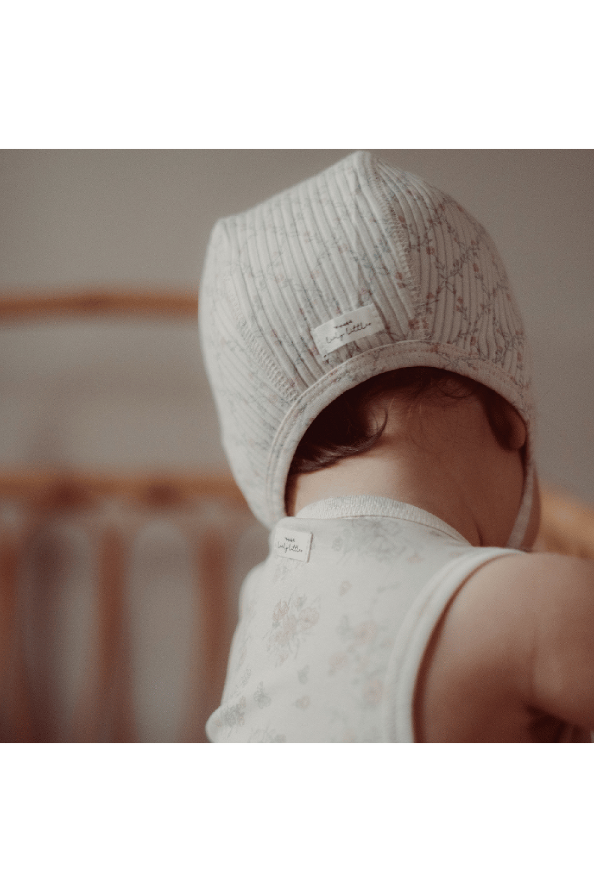 A baby wearing a light gray hat with a textured pattern and a label, sitting in front of a wooden crib.