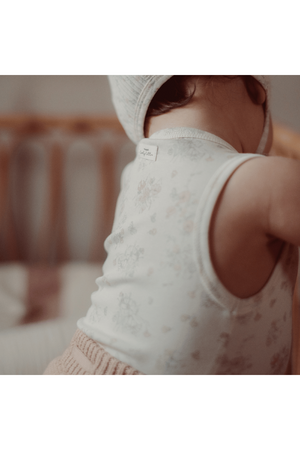 A close-up of a toddler wearing a light-colored, floral-printed cotton tank top, showcasing soft fabric and a tag on the back.
