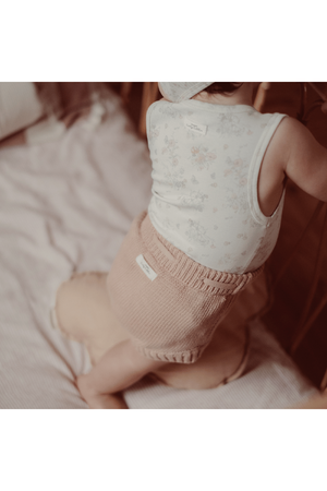 A baby wearing a floral-patterned onesie and knitted shorts sits on a soft surface, showcasing a cozy, comfortable outfit ideal for play. The setting is warm and inviting.