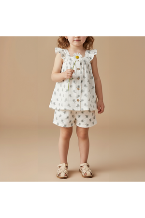 A young child stands against a beige background, wearing a white flutter top adorned with floral patterns and matching shorts. She holds a daisy, showcasing a playful summer style.