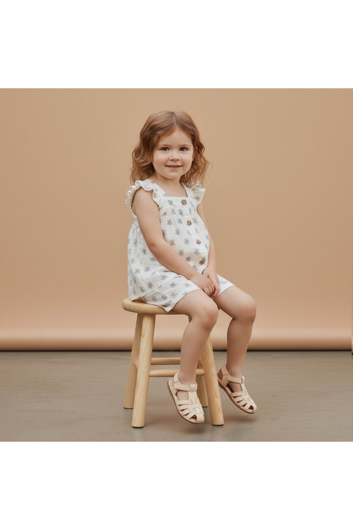 A young girl sits on a wooden stool, wearing a white organic cotton flutter top and shorts set adorned with a delicate floral pattern, against a soft beige backdrop.