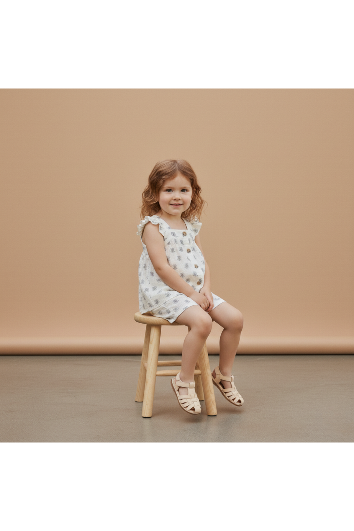 A young girl sits on a wooden stool, wearing a white organic cotton dress featuring a playful pattern. The soft background complements her relaxed summer style, ideal for playdates.
