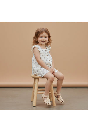 A young girl sits on a wooden stool, wearing a white organic cotton flutter top and shorts set adorned with a delicate floral pattern, against a soft beige backdrop.