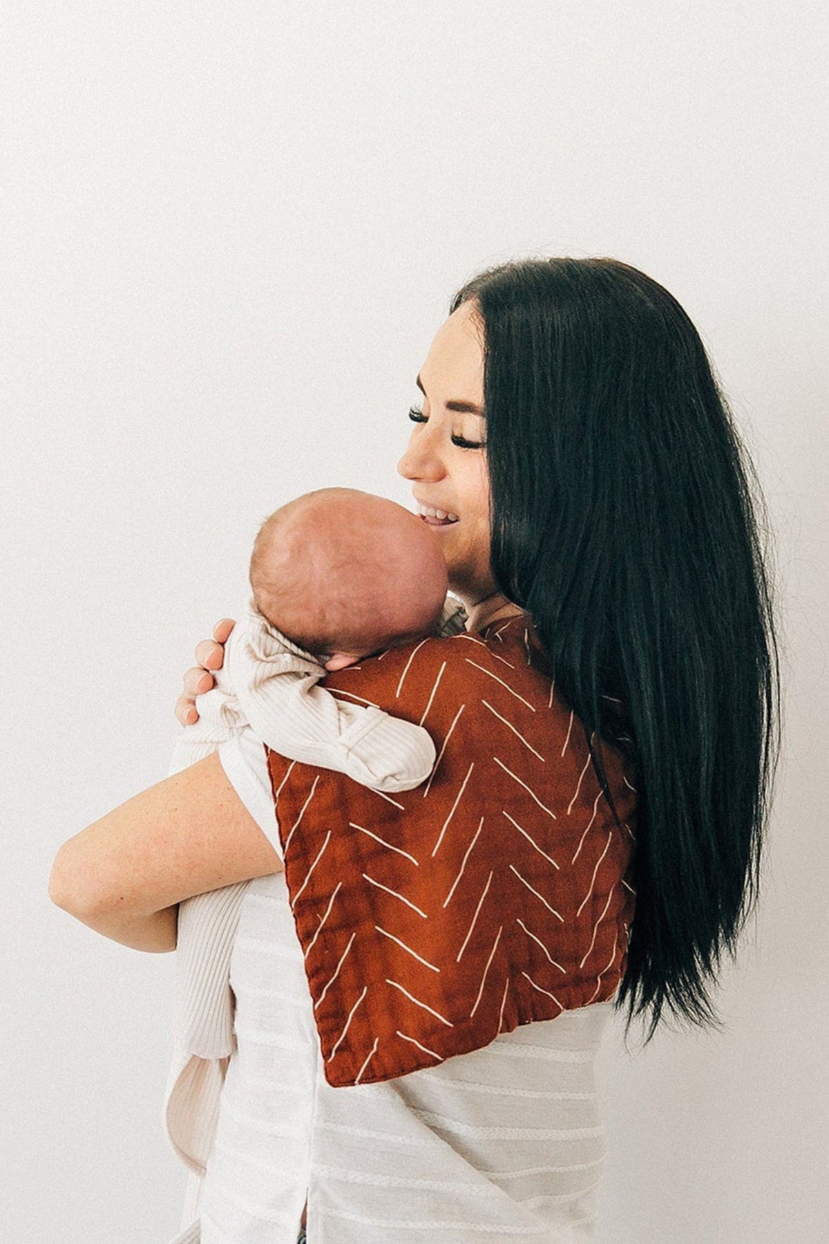 A woman holds a baby with a rust-colored, patterned burp cloth on her shoulder, smiling gently.