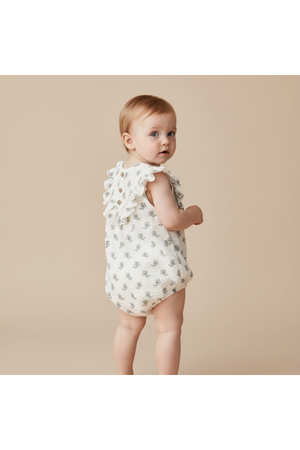 A baby girl stands facing away, wearing a sleeveless white romper adorned with whimsical patterns and ruffle details. The soft, organic cotton fabric is perfect for comfortable play.