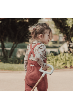 A child walks outdoors wearing rosewood knit cotton overalls paired with a floral long-sleeve top. The overalls feature adjustable straps and a cozy fit, perfect for cooler weather.