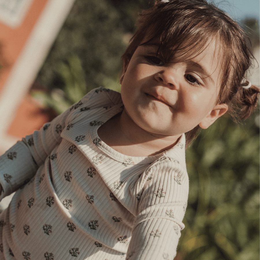 A smiling toddler with pigtails wears a light-colored long-sleeve top featuring a delicate floral print. The background shows lush greenery, creating a cheerful, outdoor setting.