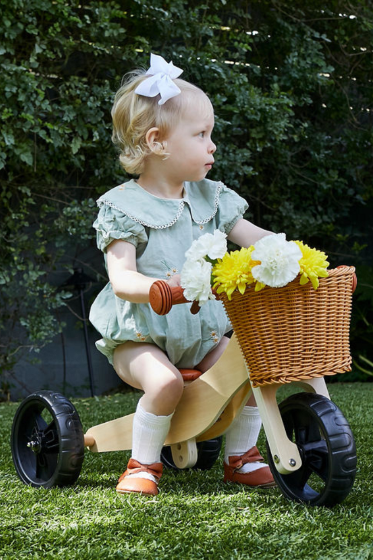 A young child sits on a wooden balance bike in a garden setting, holding a basket of flowers.