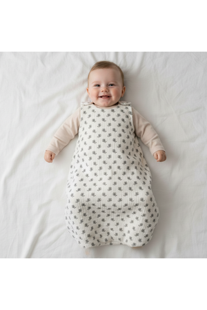 A smiling baby lies on a white blanket, wearing an organic cotton wearable blanket with a floral print in soft gray tones. The design features snap closures for easy wear.