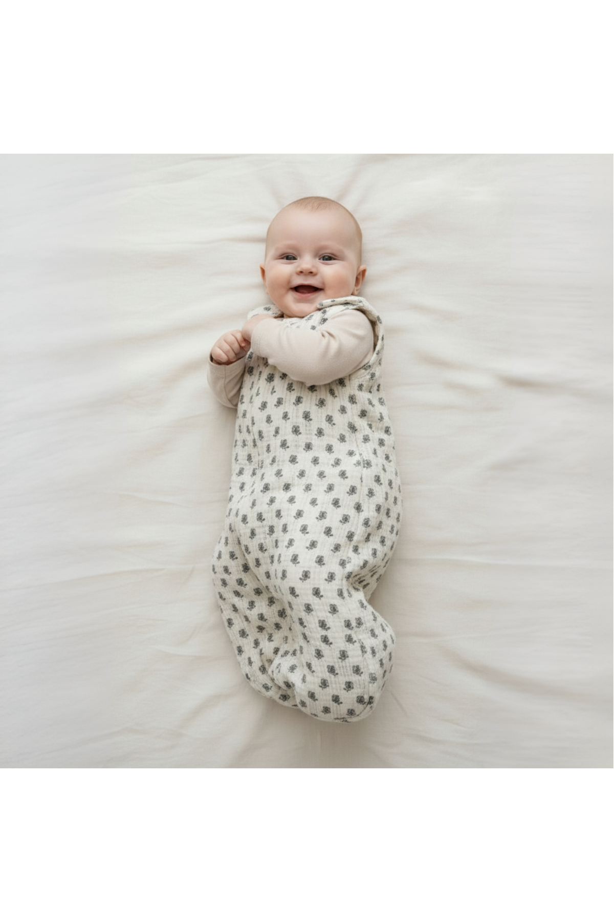 A smiling baby lies on a soft, light-colored surface, wearing a cozy, organic cotton wearable blanket with a subtle leaf pattern. The blanket ensures comfort and warmth for infants.