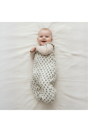 A smiling baby lies on a soft, light-colored surface, wearing a cozy, organic cotton wearable blanket with a subtle leaf pattern. The blanket ensures comfort and warmth for infants.