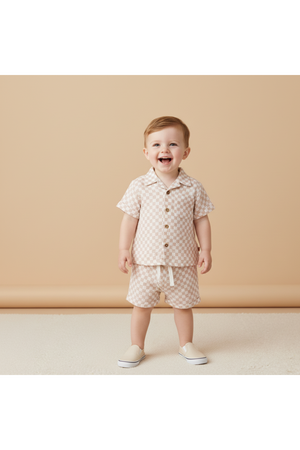 A cheerful child stands in a light beige room, wearing a brick check-patterned organic cotton shirt and shorts set, paired with white slip-on shoes, showcasing a playful summer look.