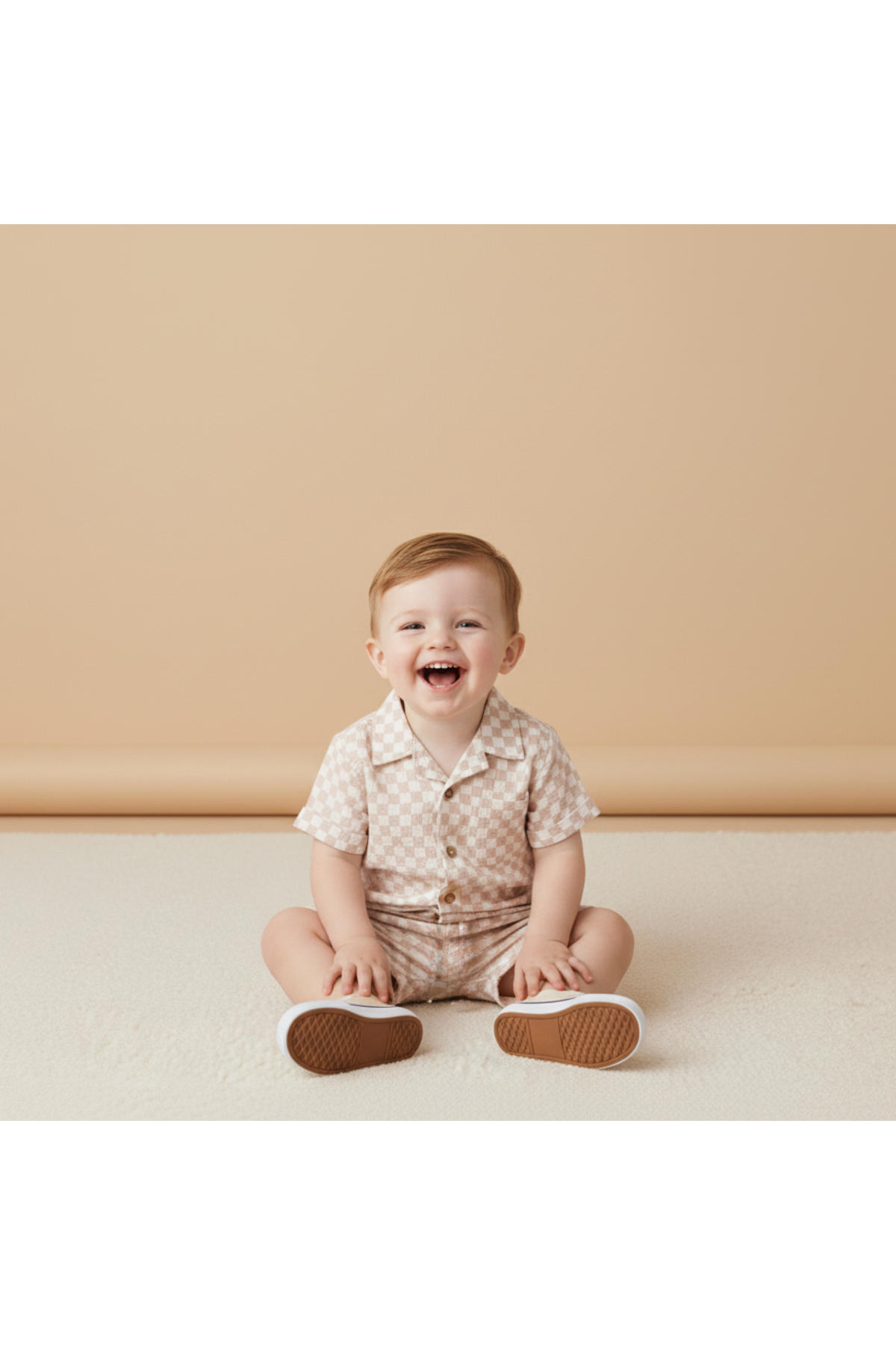 A cheerful toddler sits on a soft, light-colored surface, wearing a short-sleeve, button-up shirt and shorts set with a subtle check pattern.