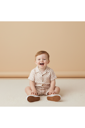 A cheerful toddler sits on a soft, light-colored surface, wearing a short-sleeve, button-up shirt and shorts set with a subtle check pattern.