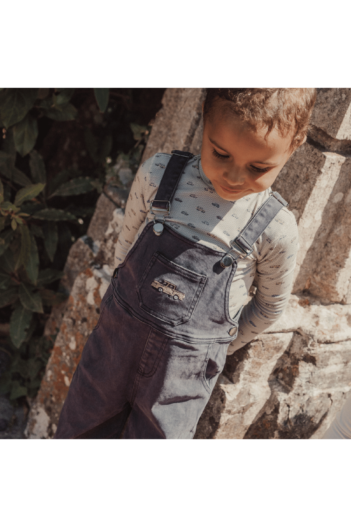 A young child wearing a light blue patterned long-sleeve shirt and dark denim overalls stands against a stone wall, surrounded by greenery, creating a charming outdoor scene.