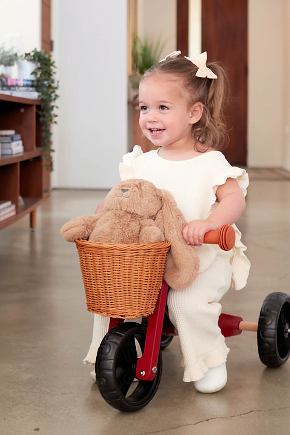 A joyful toddler rides a red balance bike with a woven basket holding a stuffed bunny. The child wears a cream outfit, showcasing a playful indoor setting with greenery.