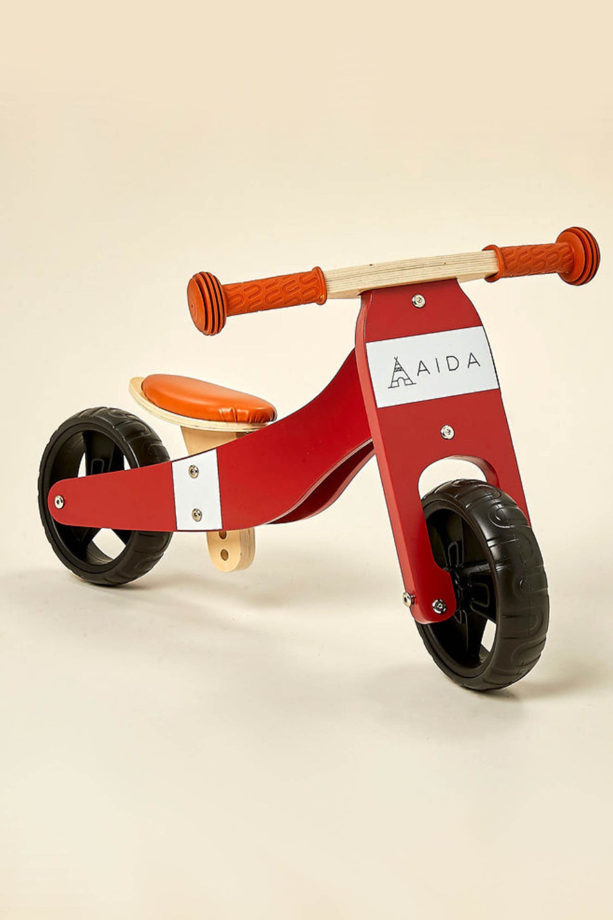 A red and white wooden balance bike with a comfortable orange seat and rubber wheels is displayed against a soft beige background.