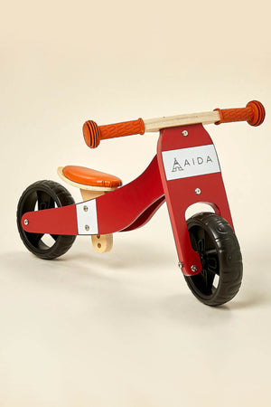 A red and white wooden balance bike with a comfortable orange seat and rubber wheels is displayed against a soft beige background.